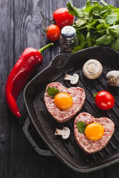 Hamburguesa corazón crudo con huevo sobre un fondo negro con verduras y hierbas . — Foto de Stock