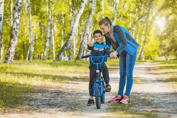 Happy kid cyclist learning to ride a bike with mom in the sunny forest on a bike — Stock Photo, Image