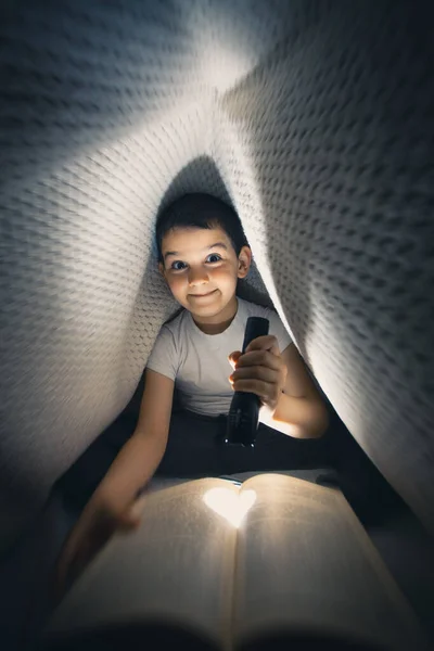 Criança feliz deitado na cama sob cobertor segurando lanterna e livro de leitura tarde da noite — Fotografia de Stock