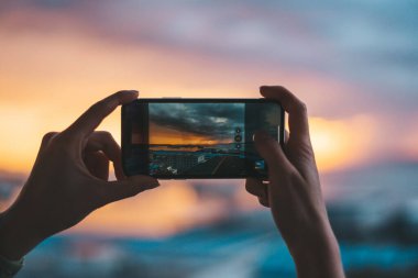 Gün batımında denizin fotoğrafını çeken akıllı bir telefon tutan eller.