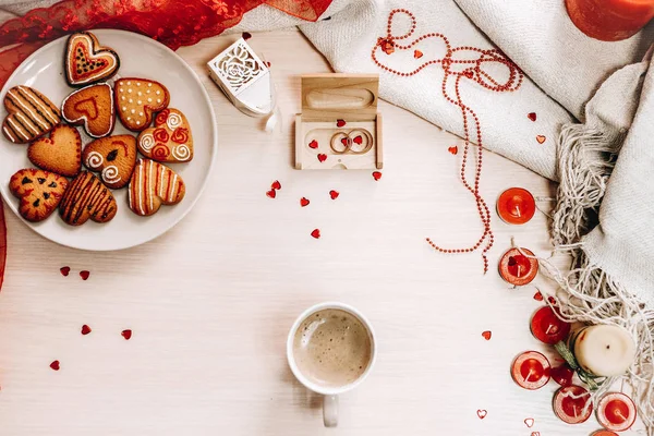 Composition with mug with beverage, decorated heart shaped cookies, red ribbon, candles, threads, golden wedding rings and red glitter hearts on background.