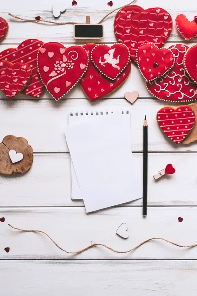 Festive composition with red glazed cookies, paper piece and pencil for romantic lettering on wooden planks background for valentines day.