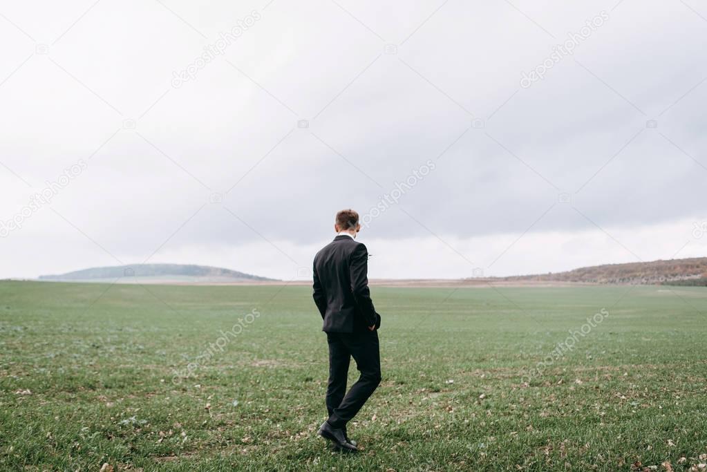 Rear view of man wearing dark evening tuxedo walking on green field