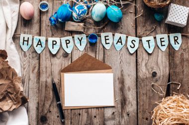 Mavi renkli yumurta, bıldırcın yumurtası, hediye kutusu, zarf açık mektup, kalem, Happy Easter yazı, saman, kumaş ve kağıt üzerinde ahşap plakalar arka plan ile mavi bayrak ile şenlikli kompozisyon Üstten Görünüm
