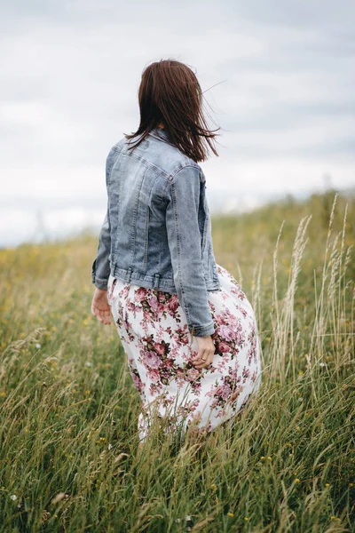 Rear view of woman in denim clothes and light skirt standing on field in windy weather