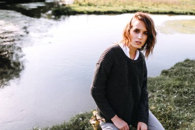 Örgü kazaklı bir kadın göletin kenarında çömelmiş kameraya bakıyor..