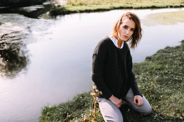 身穿针织毛衣的女人蹲在池塘边 看着相机 — 图库照片