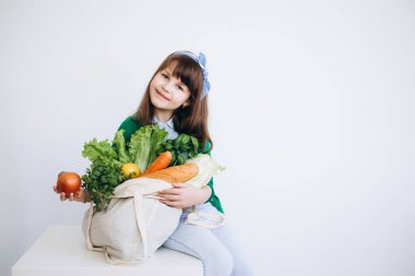 Elinde sebzeli tekstil poşeti tutan küçük bir kız. Sıfır atık. Paketsiz yiyecek alışverişi. Organik meyve ve sebzelerle dolu çevre dostu doğal çanta..