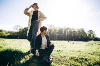 Yeşil alanda sigara içen genç adam, arka planda kadın.