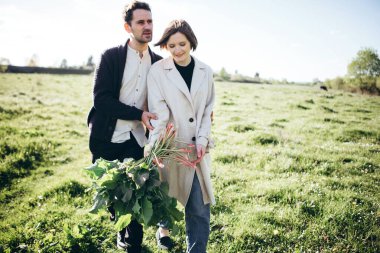Hipster çift baharda gün batımında tarlada yürüyor. Çiçek buketi tutan kadın..