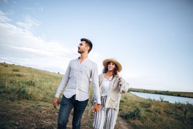 Sahada birbirine aşık mutlu çift kucaklaşıyor, öpüşüyor ve gökyüzüne karşı gülümsüyor. Şapka kızın elinde.