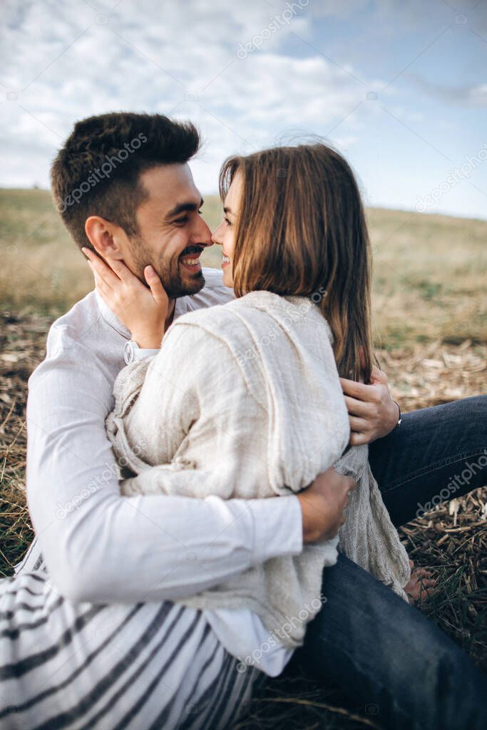 Lovely couple hugging, kissing and smiling against the sky seating on grass. Close-up portrait