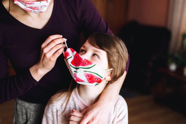 Mother puts on the daughter a mask with a bright watermelon print to protect her from the virus. Coronavirus pandemic. Stay home to stop the spread of the Corona.