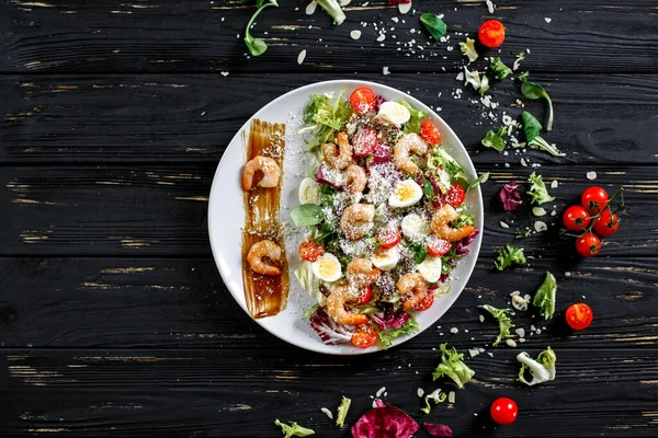 Shrimp salad on a dark background. Shrimp, lettuce, quail eggs, cherry tomatoes, parmesan, spicy sauce