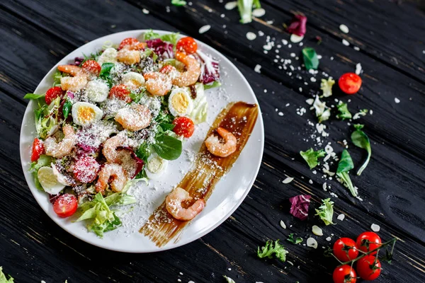 Shrimp salad on a dark background. Shrimp, lettuce, quail eggs, cherry tomatoes, parmesan, spicy sauce