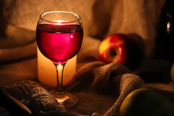 A glass full of red wine with candle muffled light shining through glass behind it and fruis on a sackcloth background. — ストック写真