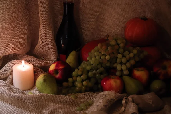 Raccolta autunnale natura morta. Mele, pere, uva e zucca su sottofondo di sacco con bottiglia di vino e lume di candela Foto Stock