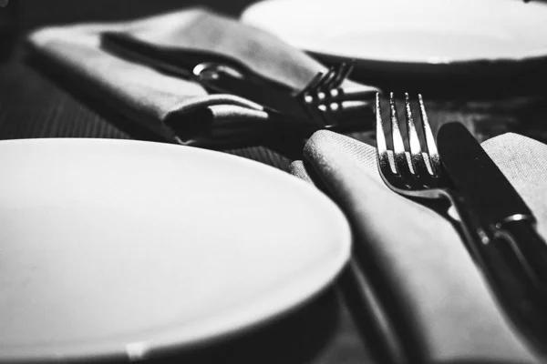 Apparecchiatura tavolo per due con piatti bianchi vuoti, forchette e coltelli su un tavolo di legno in un ristorante. Foto in bianco e nero Fotografia Stock