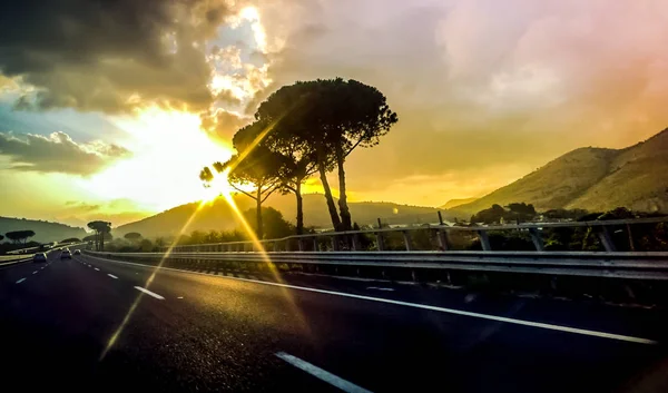 Schöne Autobahn Landschaft Blick auf Straße, Berge und Bäume auf goldenem Himmel Hintergrund mit rosa Wolken und Sonnenstrahlen — Stockfoto