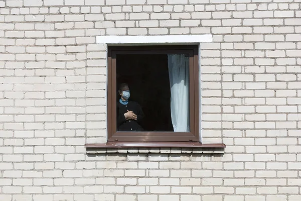 En gammal mormor i en skyddande mask tittar ut genom fönstret på självisolering. — Stockfoto