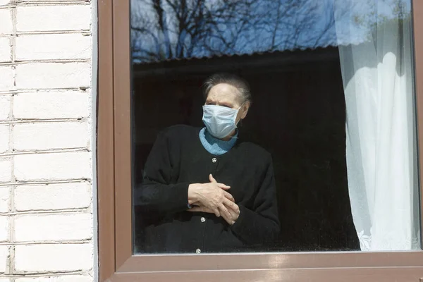 Uma velha avó em uma máscara protetora olha pela janela para o auto-isolamento . — Fotografia de Stock