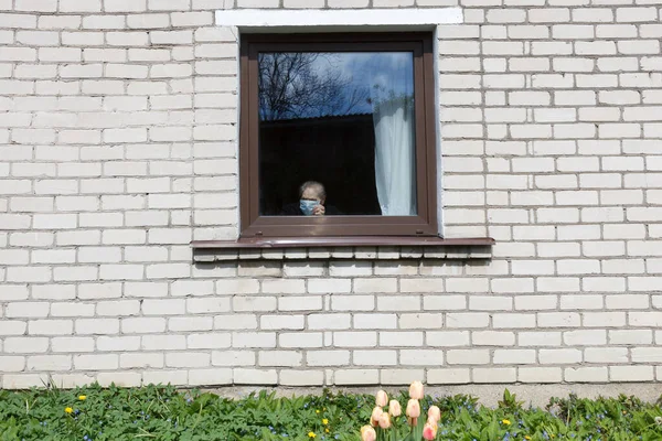 En gammal mormor i en skyddande mask tittar ut genom fönstret på självisolering. — Stockfoto