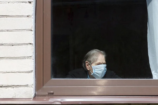 En gammal mormor i en skyddande mask tittar ut genom fönstret på självisolering. Stockbild
