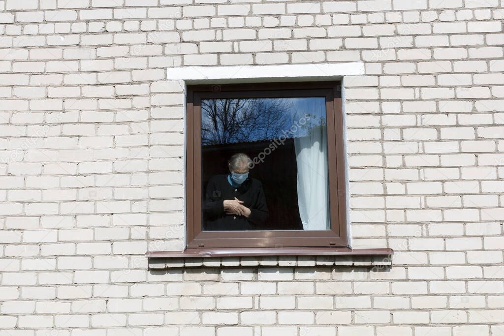 An old grandmother in a protective mask looks out the window at self-isolation.