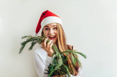 Kırmızı Noel Baba şapkalı güzel kız elinde köknar ağacı dalını tutuyor beyaz arka planda izole edilmiş mutlu ve heyecanlı görünüyor. Genç bir kadın gerçek duyguları resmediyor. Mutlu Noeller ve yeni yıl tatili