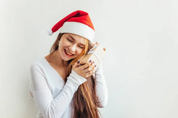 Belle fille aux cheveux longs en chapeau rouge Père Noël tenant boîte cadeau isolé sur fond blanc regardant heureux et excité. Une jeune femme dépeint de vraies émotions. Joyeux Noël et Nouvel An — Photo