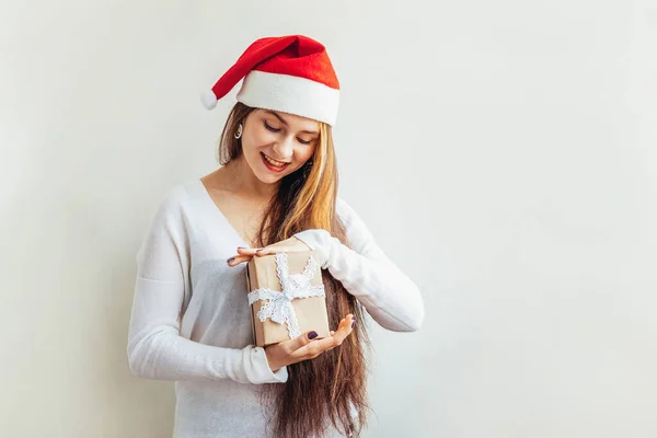 Belle fille aux cheveux longs en chapeau rouge Père Noël tenant boîte cadeau isolé sur fond blanc regardant heureux et excité. Une jeune femme dépeint de vraies émotions. Joyeux Noël et Nouvel An — Photo
