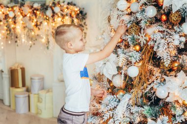 Noel arifesinde evde Noel ağacı süsleyen genç bir çocuk. Küçük çocuk kış dekorasyonlu, açık renk yatak odasında. Evde mutlu bir aile. Noel Yeni Yıl Kutlama Konsepti Zamanı