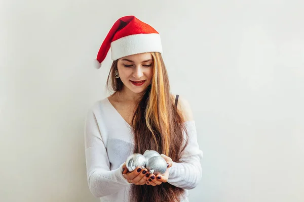 戴着红色圣诞老人帽的漂亮姑娘拿着圣诞装饰球，被白色的背景隔开，看上去既高兴又兴奋。 年轻女子刻画真实的情感。 圣诞佳节快乐 — 图库照片