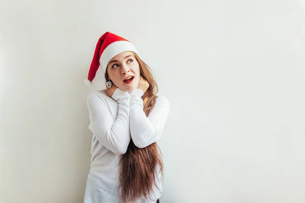 Гарна дівчина з довгим волоссям у червоному капелюсі Санта Клауса ізольована на білому фоні, виглядає щасливою і збудженою. Портрет молодої жінки, справжні емоції. Веселого Різдва і новорічних свят — стокове фото