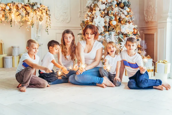 Счастливая мать семьи и пятеро детей играют в искры возле рождественской елки в канун Рождества дома. Мама, дочери, сыновья в светлой комнате с зимним убранством. Рождественский Новый Год для празднования — стоковое фото