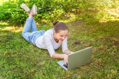 Genç kadın dizüstü bilgisayar üzerinde çalışan şehir parkında yeşil çim çim üzerinde yatan. Serbest iş konsepti