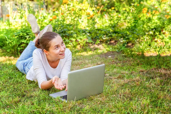 Молодая женщина, лежащая на лужайке в городском парке и работающая на ноутбуке. Концепция внештатного бизнеса — стоковое фото