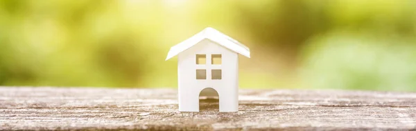 Miniature white toy model house in wooden background near green backdrop Banner