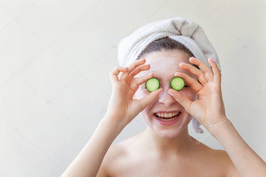 Beauty portrait of woman in towel on head with white nourishing mask or creme on face holding cucumber slices, white background isolated. Skincare cleansing eco organic cosmetic spa relax concept.