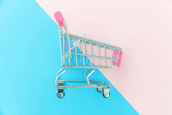 Small supermarket grocery push cart for shopping toy with wheels isolated on blue and pink pastel colorful trendy geometric background Copy space. Sale buy mall market shop consumer concept. — Stock Photo, Image