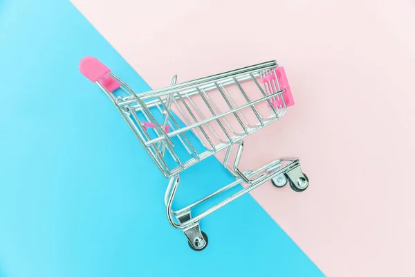 Malý nákupní vozík pro nákupní hračky s koly izolovanými na modrém a růžovém pastelovém barevném módním pozadí kopírovací prostor. Prodej koupit Obchoďák trh obchod spotřebitelský koncept. — Stock fotografie
