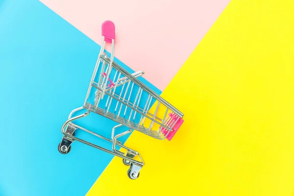 Small supermarket grocery push cart for shopping toy with wheels isolated on blue yellow pink pastel colorful trendy geometric background Copy space. Sale buy mall market shop consumer concept. — Stock Photo, Image