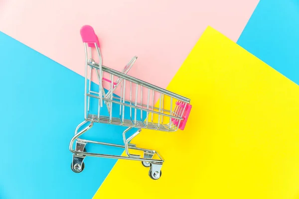 Small supermarket grocery push cart for shopping toy with wheels isolated on blue yellow pink pastel colorful trendy geometric background Copy space. Sale buy mall market shop consumer concept. — Stock Photo, Image