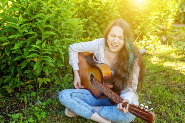 Genç hipster kadın çim oturan ve park veya bahçe arka plan üzerinde gitar çalıyor. Genç kız şarkı çalmayı öğrenme ve müzik yazma