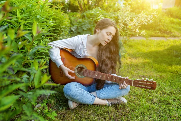 Young hipster woman sitting in grass and playing guitar on park or garden background. Teen girl learning to play song and writing music — Stock Photo, Image