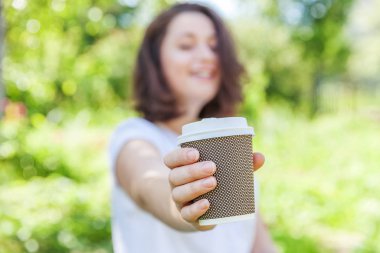 Happy girl smiling outdoor having lunch break. Beautiful young brunete woman with take away coffee cup resting on park or garden green background. Education freelancer resting leisure concept.