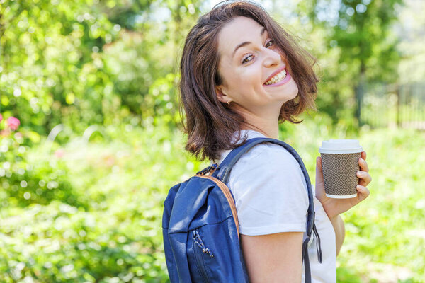 Happy positive student girl with backpack smiling on green park background. Woman with take away coffee cup having rest in campus during lunch break. Education and leisure concept