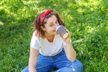 Happy girl smiling outdoor having lunch break. Beautiful young brunete woman with take away coffee cup resting on park or garden green grass background. Education freelancer resting leisure concept
