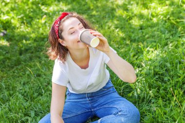 Happy girl smiling outdoor having lunch break. Beautiful young brunete woman with take away coffee cup resting on park or garden green grass background. Education freelancer resting leisure concept