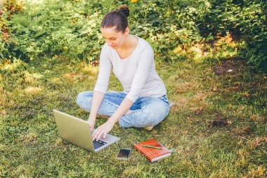 Mobil Ofis. Serbest iş konsepti. Şehir parkında yeşil çimlerde oturan genç bir kadın dizüstü bilgisayarla çalışıyor. Yaşam tarzı otantik, açık havada okuyan samimi öğrenci kız.
